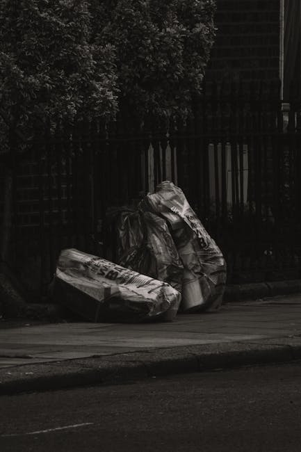 Two large black refuse bags made of plastic and possibly thick waste sack material are placed on the curbside pavement, leaning against a black wooden fence with evenly spaced vertical slats. The bags