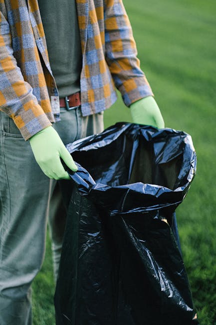 A pile of discarded cardboard boxes and large white fabric or plastic bags filled with waste materials are situated on the ground next to a rough brick wall and a concrete block wall, partially shaded
